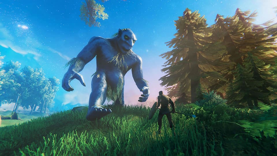 An in-game screenshot from Valheim showing a massive blue troll towering over a player equipped with a shield and spear. The setting is a lush, grassy area surrounded by tall pine trees under a bright, starry sky.